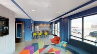 This energetic single-floor space makes an immediate impression with its bold cobalt blue walls, crisp white ceiling, and polished concrete floors. The open-plan layout seamlessly connects a casual lounge area with a functional kitchenette and dining zone, creating a versatile environment suited for both relaxation and social interaction.

The lounge area is anchored by a lively geometric-patterned area rug in multicolored tones, complemented by a mix of modular seating including colorful hexagonal ottomans and a vibrant orange accent chair paired with a teal side table. A sleek dark sofa adds a grounding contrast to the playful palette.

Adjacent to the lounge, the dining area features a modern high-top table surrounded by bright yellow chairs, contributing to the space's energetic aesthetic. The kitchenette behind it is equipped with a full-height refrigerator, countertop appliances, and ample counter space, making it practical for everyday use.

Large floor-to-ceiling windows and glass doors along one wall flood the interior with natural light while offering views of the exterior surroundings. Recessed ceiling lighting provides consistent, even illumination throughout. The overall ambiance is modern, lively, and welcoming — thoughtfully designed to foster a dynamic and community-oriented atmosphere.