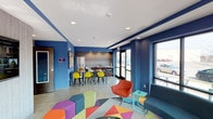 This energetic single-floor space makes an immediate impression with its bold cobalt blue walls, crisp white ceiling, and polished concrete floors. The open-plan layout seamlessly connects a casual lounge area with a functional kitchenette and dining zone, creating a versatile environment suited for both relaxation and social interaction.

The lounge area is anchored by a lively geometric-patterned area rug in multicolored tones, complemented by a mix of modular seating including colorful hexagonal ottomans and a vibrant orange accent chair paired with a teal side table. A sleek dark sofa adds a grounding contrast to the playful palette.

Adjacent to the lounge, the dining area features a modern high-top table surrounded by bright yellow chairs, contributing to the space's energetic aesthetic. The kitchenette behind it is equipped with a full-height refrigerator, countertop appliances, and ample counter space, making it practical for everyday use.

Large floor-to-ceiling windows and glass doors along one wall flood the interior with natural light while offering views of the exterior surroundings. Recessed ceiling lighting provides consistent, even illumination throughout. The overall ambiance is modern, lively, and welcoming — thoughtfully designed to foster a dynamic and community-oriented atmosphere.