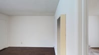 This single-floor space presents a straightforward and functional interior defined by its clean, minimalist aesthetic. The walls are finished in crisp white, creating a bright and open atmosphere that allows for versatile use of the space. Underfoot, rich dark hardwood-style flooring adds warmth and contrast against the light walls, grounding the interior with a classic, practical finish. The ceiling features a textured popcorn finish, a detail that adds subtle depth to the otherwise smooth surfaces throughout. A prominent built-in HVAC wall unit is positioned along one wall, indicating dedicated climate control for year-round comfort. The overall layout is open and unobstructed, providing a blank canvas well-suited for a variety of configurations. The space is compact yet functional, with clean lines and neutral finishes that make it adaptable to different uses or furnishing styles. The combination of white walls, dark flooring, and the textured ceiling gives the interior a no-frills, utilitarian character that prioritizes practicality and ease of maintenance.
