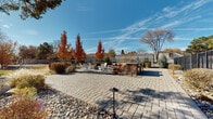 This thoughtfully landscaped outdoor space offers a welcoming and functional environment centered around a generously sized cobblestone paver patio. The hardscape serves as the primary gathering area and is complemented by a handsome stacked-stone fire pit, around which a cluster of white Adirondack chairs is arranged — ideal for relaxed outdoor socializing. Decorative low-voltage landscape lighting is integrated throughout the patio area, providing subtle illumination after dark.

Surrounding the paved area, the grounds feature a combination of open lawn, river rock mulch beds, ornamental grasses, and mixed shrubbery that provide visual interest across all seasons. Standout landscape elements include a central planting island with vibrantly colored autumn maples, dwarf conifers, and rounded shrubs that add structure and seasonal color to the space. Mature deciduous and evergreen trees border the perimeter, offering both shade and a sense of enclosure.

The property is fully enclosed by a black wrought-iron style fence, providing both security and an open, airy feel. A tennis court is visible just beyond the fencing on one side, adding to the recreational amenities of the space. The overall ambiance is bright, open, and inviting, with expansive views of a clear blue sky and a well-maintained, naturalistic landscape design throughout.