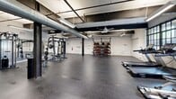 Located at 1700 Fairview St in Greensboro, NC, this fitness facility offers a spacious, single-floor exercise room with a distinctive industrial aesthetic. The open layout is defined by exposed steel beams, large-diameter ventilation ductwork running along the ceiling, and white-painted brick walls that lend a raw, warehouse-inspired character to the space. Oversized steel-framed windows line the walls, flooding the interior with abundant natural light and providing views of the surrounding greenery. Ceiling fans are strategically placed throughout to ensure comfortable airflow during workouts.

The facility is comprehensively equipped to support a wide range of fitness activities. The cardio zone features multiple treadmills, elliptical trainers, and stationary and cycling bikes arranged along the windowed walls. A dedicated strength training area includes a power rack with free weights, a cable machine, dumbbell racks, and a medicine ball storage unit. The rubber-floored open floor space provides ample room for functional training and stretching. Additional amenities include dedicated restroom facilities accessible directly from the workout floor. The combination of high ceilings, natural light, and thoughtfully arranged equipment creates an energizing and functional environment well-suited for comprehensive fitness routines.