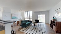 This contemporary two-story space is defined by its striking architectural design and open, airy atmosphere. The main living area immediately impresses with soaring ceilings that amplify the sense of volume throughout the room. A dramatic wall of floor-to-ceiling windows floods the space with natural light while framing expansive urban views, including a notable stadium or bridge structure visible in the distance — a standout feature that adds character and a strong sense of place.

The living area is spacious and versatile, featuring light hardwood flooring that runs throughout and contributes to a warm, modern aesthetic. A sliding glass door provides direct access to an outdoor area, further enhancing the connection between interior and exterior spaces. The clean white walls serve as a neutral backdrop, complementing the open layout and allowing the architectural details to take center stage.

A staircase leads to the second floor, adding functional depth to the overall layout. The space's design balances minimalism with warmth, making it well-suited for both everyday living and entertaining. Track lighting adds a practical yet stylish element to the ceiling, while the generous proportions of the main level ensure a comfortable and inviting environment throughout.