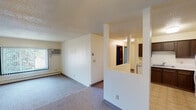 Located at 418 W Apollo Ave in Bismarck, North Dakota, this single-floor unit offers a straightforward and functional layout well-suited for comfortable everyday living. The spacious living area is finished with wall-to-wall carpeting, a ceiling fan with light fixture, and a large window dressed with vertical blinds that allows natural light to flow in while offering views of surrounding greenery. A wall-unit air conditioner and baseboard heating ensure year-round climate control. A half-wall partition divides the living space from the kitchen, creating an open yet defined layout. The kitchen features dark wood cabinetry with ample upper and lower storage, light-toned countertops, a double-basin sink, a white dishwasher, and tile flooring that contrasts with the carpeted living areas. A hallway leads from the main living space to the bedroom and bathroom. The bedroom is a generously sized, carpeted room with neutral walls, a textured ceiling, and baseboard heating. The full bathroom is accessed through dark wood doors and includes a tiled tub-and-shower combination, a toilet, a vanity with dark cabinetry, and tile flooring consistent with the kitchen. The textured ceilings appear throughout the unit, contributing to a cohesive interior character.