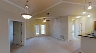 This single-story space showcases a spacious, open main living area defined by vaulted ceilings that create an airy, expansive feel. The ceiling transitions from a raised center section to angled slopes on either side, adding architectural interest and visual depth to the room. Neutral beige walls and light-colored carpeting throughout contribute to a clean, understated aesthetic that complements a variety of furnishing styles.

A decorative pendant light hangs near the entry, providing a focal point upon arrival, while a ceiling fan with a light fixture is centrally positioned in the main living area, offering both functional airflow and ambient lighting. French doors with glass pane detailing allow natural light to filter into the space, enhancing its open and welcoming atmosphere.

Flush with the wall, HVAC vents are positioned at both floor and ceiling levels, ensuring efficient climate control throughout. A doorway on the far side of the main room leads to an additional area, suggesting further functional spaces beyond the primary living zone. White trim and baseboards provide clean contrast against the warm wall tones, completing the space's cohesive, well-maintained interior.