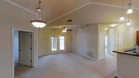 This single-story space showcases a spacious, open main living area defined by vaulted ceilings that create an airy, expansive feel. The ceiling transitions from a raised center section to angled slopes on either side, adding architectural interest and visual depth to the room. Neutral beige walls and light-colored carpeting throughout contribute to a clean, understated aesthetic that complements a variety of furnishing styles.

A decorative pendant light hangs near the entry, providing a focal point upon arrival, while a ceiling fan with a light fixture is centrally positioned in the main living area, offering both functional airflow and ambient lighting. French doors with glass pane detailing allow natural light to filter into the space, enhancing its open and welcoming atmosphere.

Flush with the wall, HVAC vents are positioned at both floor and ceiling levels, ensuring efficient climate control throughout. A doorway on the far side of the main room leads to an additional area, suggesting further functional spaces beyond the primary living zone. White trim and baseboards provide clean contrast against the warm wall tones, completing the space's cohesive, well-maintained interior.