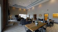 This single-floor space showcases a thoughtfully designed open-concept layout that immediately impresses with its soaring tray ceilings, accented with layered crown molding in a contrasting gray tone. Recessed lighting is strategically placed throughout, ensuring the space is well-lit and inviting at all times of day. Large windows along the far wall allow natural light to pour in, enhancing the airy, expansive atmosphere.

The main gathering area is anchored by a rustic reclaimed wood communal table paired with industrial-style metal chairs, creating a casual yet functional focal point. Adjacent to this, a comfortable lounge zone features plush dark sofas and accent chairs arranged around a low-profile coffee table, ideal for relaxed conversation or informal meetings. A large-screen television is mounted nearby, adding to the room's versatile functionality.

Warm wood-tone French doors lead to additional areas, while decorative touches such as an oversized wall clock and potted greenery add character and charm. The light tile flooring complements the neutral gray-and-white color palette, tying the entire space together. Overall, this space combines modern design sensibility with welcoming, functional furnishings to create a polished and comfortable communal environment.