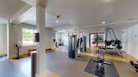 This expansive fitness center is thoughtfully designed across a single floor, offering a comprehensive range of exercise zones to accommodate diverse workout needs. The facility is bathed in natural light from numerous windows that frame views of the surrounding greenery, creating an open and energizing atmosphere throughout.

The cardio zone is prominently featured along one wall, lined with multiple treadmills and elliptical trainers, complemented by a large floor-to-ceiling mirror that visually expands the space. Adjacent areas house stationary cycling bikes equipped with individual screens, as well as rowing machines for full-body conditioning.

Strength training is well represented through a dedicated zone featuring a variety of cable and resistance machines, including multi-function cable towers and cable-guided press machines. Free-standing columns divide the space into functional workout areas without compromising the overall sense of openness.

The flooring transitions between hardwood-style surfaces, rubberized grey mats, and vibrant yellow-accented zones, clearly delineating different activity areas. Recessed lighting supplements the abundant natural light, while a wall clock and wall-mounted displays add practical touches. A separate, enclosed room with glass partition walls provides additional workout space, housing medicine balls, resistance equipment, and more. The facility also includes a dedicated exit door for convenient access.