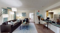 This single-floor residence offers a thoughtfully designed layout that balances comfort and functionality across multiple rooms. The entry hallway, finished with rich hardwood-style flooring, leads seamlessly into the main living areas. The living room is a welcoming space featuring plush carpeting, a textured ceiling, and generous natural light streaming through large windows dressed with teal curtains. A glass-top coffee table and ample seating area complete the room, which also provides direct access to an exterior door. Adjacent to the living room, a pass-through connects to the dining and kitchen area. The kitchen is well-equipped with warm wood cabinetry, dark granite-style countertops, stainless steel appliances including a range and refrigerator, and hardwood-style flooring. A dedicated laundry area is accessible directly from the kitchen. The unit includes multiple bedrooms, each carpeted and bright, with windows allowing natural light. Two full bathrooms are present, each featuring hardwood-style flooring, granite-topped vanities with single-basin sinks, large mirrors with overhead lighting, and a shower-tub combination. The overall ambiance is bright, clean, and livable, with a consistent design palette throughout.
