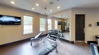 This fitness center is designed to provide a comprehensive workout experience within a thoughtfully organized, single-floor layout. The space features durable wood-look flooring throughout and is illuminated by a combination of recessed lighting and decorative pendant ceiling fans, creating a bright and energizing environment.

The strength training area is the centerpiece of the facility, housing a multi-station cable machine, a squat rack with weight plates, and a full dumbbell rack with an adjustable bench for targeted lifting sessions. Floor-to-ceiling mirrors line the walls, amplifying the sense of space and allowing users to monitor their form during workouts. Additional weight benches are positioned throughout for versatile training options.

Natural light filters in through multiple windows, offering views of the surrounding greenery and adding an open, airy quality to the workout environment. A wall-mounted television provides entertainment during exercise sessions.

The lobby and entry area is clean and functional, featuring navy blue cabinetry with a compact refrigerator, a wall-mounted television, and informational displays — offering a welcoming transition point into the facility. Dark-framed doors provide stylish access between zones. The overall design strikes a balance between practicality and polished aesthetics, making this fitness center a well-rounded space for a variety of training routines.