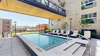Located in the heart of Salt Lake City at 360 W 200 S, this outdoor amenity deck delivers a polished, resort-inspired experience designed for relaxation and social gathering. At its centerpiece sits a sparkling pool complemented by an adjacent spa, both framed by clean tile surrounds and accessed via stainless steel handrails. A striking wood-paneled overhead canopy extends above the pool area, providing partial shade while preserving open views of the surrounding cityscape and neighboring historic brick buildings.

A generous sun deck lines the pool, furnished with rows of white chaise lounges and accent tables, ideal for enjoying Utah's abundant sunshine. Lush ornamental grasses, young trees, and curated planters are woven throughout the space, softening the modern architectural palette of light-toned panels and dark metal railings.

Beyond the pool zone, a spacious courtyard unfolds, featuring multiple distinct seating areas with stylish outdoor furniture, including woven lounge chairs, dining tables, and casual benches. String lights are strung overhead, creating an inviting ambiance for evening gatherings. Wide-format tile paving, accessible ramps, and thoughtful greenery integrate seamlessly to create a cohesive, well-appointed outdoor retreat in the center of downtown Salt Lake City.
