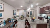 This single-story residence is designed with an open-concept layout that seamlessly connects the kitchen, dining, and living areas, creating a cohesive and functional living environment. The kitchen is a standout feature, showcasing rich dark-wood cabinetry, granite countertops, a subway tile backsplash, and a full suite of stainless steel appliances including a range, microwave, and refrigerator. A large granite-topped island with pendant lighting overhead serves as both a prep space and casual dining area with bar seating.

The dining area sits adjacent to the kitchen, flowing naturally into the living room, which features light wood-style flooring, crown molding, a ceiling fan, and a large window dressed with patterned curtains that allows natural light to fill the space. The entryway is clean and welcoming, accented with crown molding and decorative wall art.

The bedroom is carpeted for comfort and includes a window with blinds and teal drape panels, offering natural light and privacy. It connects to a full bathroom finished with tile flooring, a vanity with dark cabinetry, and a shower. A walk-in closet with built-in shelving provides ample storage. The overall aesthetic is modern and cohesive throughout.