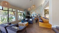 Located at 10757 Lemon Ave in Rancho Cucamonga, CA, this stylish single-story community clubhouse offers a thoughtfully designed gathering space with a contemporary aesthetic. The expansive main lounge features soaring vaulted ceilings, elegant herringbone-pattern hardwood flooring, and an abundance of natural light streaming through large floor-to-ceiling windows overlooking the outdoor pool area. A dramatic floor-to-ceiling fireplace clad in sleek concrete serves as the focal point of the lounge, framed by a wall-mounted TV and complemented by plush tufted sofas, live-edge wood coffee tables, and woven rattan pendant lighting that adds warmth to the space. A built-in seating platform with cushions and a dedicated workstation area make this lounge as functional as it is inviting. The kitchenette area is appointed with soft blue cabinetry, white quartz countertops, and a stainless steel sink, seamlessly connected to a cozy dining nook. The dining area features a round wood table and chairs set beside roller shade windows and glass doors that open directly to the pool deck, blending indoor and outdoor living. Recessed lighting and carefully curated artwork and décor complete the polished, resort-inspired ambiance throughout.
