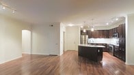 This single-floor space offers a well-designed open-concept layout that seamlessly connects the living area and kitchen, creating a spacious and cohesive environment. Rich, dark hardwood flooring runs throughout the main living areas, adding warmth and a polished aesthetic to the interior.

The kitchen is a standout feature, showcasing deep espresso-toned cabinetry that extends to the upper walls, paired with light countertops that provide a striking contrast. A curved kitchen island with an integrated sink serves as a central focal point, offering both functional workspace and a visually dynamic element. Stainless steel appliances, including a full-sized refrigerator, contribute a modern, professional feel. Elegant pendant lighting hangs above the island, further enhancing the kitchen's refined character.

The adjacent living area is open and versatile, offering ample room for a variety of furniture arrangements. Neutral, light-toned walls and white trim work complement the flooring and cabinetry throughout, maintaining a clean and classic palette. Crown molding adds an architectural detail that elevates the overall finish of the space. Multiple doorways provide access to additional rooms, suggesting a practical and well-connected floor plan. The overall ambiance is contemporary yet inviting, with a strong emphasis on quality finishes and thoughtful design.