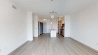 This single-floor unit showcases a clean, contemporary design with an open-concept layout that seamlessly integrates the living area and kitchen. Light hardwood-style flooring extends throughout the space, contributing to a bright and cohesive aesthetic. The crisp white walls and smooth ceilings are complemented by tasteful track lighting, which provides both functional illumination and a modern accent to the interior.

The kitchen is a standout feature, equipped with stainless steel appliances including a refrigerator, microwave, and stove. Warm wood-toned cabinetry lines the walls, offering ample storage, while a central white island with an integrated sink serves as a practical and stylish focal point. The open-concept design ensures the kitchen flows naturally into the living area, making the space feel expansive and well-connected.

The entry area is visible toward the back, featuring a neutral-toned door and a closet, providing functional storage upon arrival. The overall ambiance of the unit is modern and minimalist, with a neutral color palette that creates a clean, airy atmosphere well-suited to a variety of interior styles.