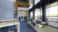 This single-floor space showcases a contemporary design aesthetic, combining industrial and natural elements to create a stylish, multifunctional lounge environment. The most commanding feature is the dramatic double-height ceiling, which amplifies the sense of openness and grandeur throughout the interior.

A textured stacked-stone accent wall serves as a bold focal point, framed by warm pendant lighting that adds an intimate ambiance to the surrounding area. Overhead, exposed wooden beam detailing introduces an organic contrast against the otherwise sleek, dark palette, lending visual warmth to the modern design.

Floor-to-ceiling windows line one side of the space, flooding the interior with natural light and offering open views of the outdoors. The flooring transitions between dark wood-style planks and area rugs, subtly defining different seating zones.

Furnishings are thoughtfully arranged to support a variety of uses. Plush gray velvet lounge chairs and metallic accent tables create intimate conversation areas, while bar-height seating near the accent wall accommodates more structured gatherings. Light wood communal tables along the perimeter provide additional flexible workspace. The overall ambiance is polished, inviting, and well-suited for socializing or quiet productivity.
