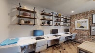 This single-floor coworking space masterfully combines industrial-modern design elements to create a functional and visually engaging work environment. The space is divided into two distinct zones that complement each other seamlessly.

The workstation area features a long, continuous white desk surface accommodating multiple users side by side, each station equipped with a monitor and ergonomic rolling chair. Above the desk, tiered open shelving constructed from reclaimed wood planks and black iron pipe brackets lines the wall, providing both storage and decorative display space for books, plants, and decorative objects. The warm hardwood flooring runs throughout, anchoring the industrial aesthetic.

Adjacent to the workstation zone is a collaborative meeting area centered around a solid wood farmhouse-style table surrounded by dark leather chairs. A striking exposed brick accent wall serves as a dramatic backdrop, adorned with framed artwork. A ceiling-mounted wooden beam installation suspends an array of Edison bulb pendant lights, casting warm ambient light across the space. A window with dark-trimmed blinds allows natural light to filter in, while a bold set of black double doors adds a refined architectural focal point. The textured ceiling and recessed lighting complete the well-layered, purposeful design of this productive shared workspace.