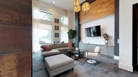 This thoughtfully designed single-floor amenity space combines contemporary aesthetics with functional comfort across several distinct areas. The main lounge commands attention with its soaring double-height ceilings and floor-to-ceiling windows dressed in sheer drapery, flooding the room with natural light and offering views of the surrounding greenery. A generous sectional sofa anchored by a patterned area rug anchors the seating area, complemented by a statement wall clad in warm wood paneling and a mounted flat-screen TV above a sleek built-in media credenza. Sculptural gold pendant lights add a refined, decorative touch overhead. A cozy bistro-style table and chairs near the windows provide an additional gathering spot. The wet bar area is a standout feature, showcasing an expansive granite-topped island with leather bar seating, iridescent mosaic tile backsplash walls in hues of gold and purple, crystal pendant lighting, and integrated appliances. Adjacent cabinetry in muted grey offers ample storage. A glass-walled fitness room, accessible through steel-framed doors, maintains visual connectivity with the rest of the space while housing exercise equipment on light hardwood flooring. Throughout, the design balances bold accents with neutral tones, creating a polished and inviting environment.
