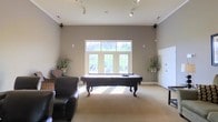 Located at Cape Landing in Myrtle Beach, South Carolina, this single-floor amenity space is designed to offer residents a comfortable and inviting gathering environment. The centerpiece of the space is a generously sized game room featuring a full-size billiard table set beneath soaring ceilings that amplify the open, airy feel of the room. Tall windows and French doors line the far wall, flooding the interior with abundant natural light while framing views of the surrounding greenery outside. Track lighting installed along the ceiling provides additional illumination, ensuring the space is well-lit at all hours. Neutral-toned walls and wall-to-wall carpeting create a warm, understated backdrop that complements the room's leisure-focused furnishings, including cushioned lounge chairs positioned for relaxed socializing. Decorative potted plants flank the windows, adding a touch of organic warmth to the space. The overall ambiance is welcoming and functional, thoughtfully designed to serve as a recreational hub where residents can unwind, socialize, and enjoy community amenities in a bright and well-appointed setting.