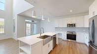 This thoughtfully designed two-story home in Stafford, TX, offers 2 bedrooms and 3 bathrooms across two well-proportioned floors. The main level features a bright, open-concept kitchen and living area with soaring double-height ceilings and an abundance of natural light streaming through multiple windows. The kitchen is a standout, showcasing a large center island with a farmhouse-style stainless steel sink, white shaker cabinetry, a light gray subway tile backsplash, pendant lighting, and a full suite of stainless steel appliances. Light hardwood flooring flows seamlessly throughout both levels, complementing the clean, neutral color palette. The living area benefits from floor-to-ceiling windows that fill the space with natural light and provide outdoor views. A convenient powder room on the main level features a modern pedestal sink with an LED-backlit mirror. Upstairs, the generously sized primary bedroom includes a ceiling fan and a walk-in closet with built-in shelving. The second bedroom is equally well-appointed with its own ceiling fan and natural light. The bathrooms are finished with marble-style tiled tub-and-shower surrounds, white vanities, and brushed nickel fixtures. Two dedicated closets add practical storage throughout the home.