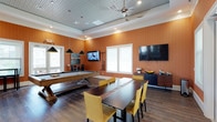 This single-floor recreational space is designed as a versatile social lounge, blending entertainment and gathering functionality in a cohesive, contemporary setting. The room is defined by warm terracotta-toned walls that create an inviting, energetic ambiance, complemented by rich hardwood flooring and crisp white crown molding that adds a polished finish.

At the heart of the space sits a full-size billiards table, positioned beneath a pair of sleek black pendant lights that provide focused task lighting. Adjacent to the billiards area, a substantial live-edge dining table surrounded by mustard-yellow upholstered chairs offers a comfortable spot for socializing or casual gatherings. Two large flat-screen TVs are mounted on the walls, ensuring excellent sightlines from multiple areas of the room.

Natural light filters in through white-trimmed windows fitted with privacy blinds, brightening the space while maintaining a cozy atmosphere. A ceiling fan overhead aids in air circulation, adding to the room's comfort. A stylish credenza along one wall provides functional storage and display space, accessorized with decorative accents. The paneled ceiling detail adds subtle architectural character, rounding out a well-appointed space built for leisure and community engagement.