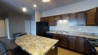 This single-floor space centers around a thoughtfully designed kitchen that serves as its standout feature. Rich, dark-stained cabinetry lines the walls, providing ample storage while lending a warm, classic aesthetic to the space. The countertops throughout the kitchen are finished in granite, displaying a natural pattern of gold, cream, and gray tones that add visual depth and durability. A large kitchen island with a matching granite surface anchors the space, offering additional workspace and casual seating potential. Black appliances, including a full-sized refrigerator and dishwasher, complement the dark cabinetry for a cohesive look. Under-cabinet lighting illuminates the work surfaces, while recessed ceiling lights and a pendant fixture provide layered illumination throughout the kitchen. A double-basin sink is integrated into the perimeter countertop for practicality. Light hardwood flooring runs throughout, creating a warm contrast to the darker upper elements. The neutral gray walls keep the overall palette clean and versatile. A white paneled door visible in the background hints at additional rooms or storage within the unit.