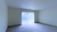 This single-floor space offers a clean, straightforward layout designed for comfortable everyday living. The main living area is a notable highlight, featuring large sliding glass doors dressed with vertical blinds that allow an abundance of natural light to fill the room while offering a view of the lush greenery outside. The soft, neutral-toned carpeting underfoot adds warmth and comfort to the space, complementing the light-colored walls that contribute to the room's bright and airy atmosphere. Overhead, the smooth ceiling hosts integrated ventilation, reflecting a practical and functional design approach. The understated, neutral palette throughout creates a versatile backdrop well-suited for a variety of interior styles and furnishing choices. The sliding glass doors not only enhance natural lighting but also provide a seamless visual connection to the outdoor environment, bringing a sense of openness to the interior. Overall, this space presents a calm and livable environment with key features that balance simplicity, natural light, and functional design.