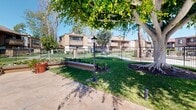 Located at 15123 Brookhurst St in Westminster, California, this outdoor community amenity space offers a well-appointed recreational environment designed for relaxation and leisure. At the heart of the space is a generously sized swimming pool with crystal-clear turquoise water, accessible via entry steps and framed by a wide concrete sun deck. The deck is furnished with lounge chairs and seating areas, providing ample room for sunbathing and socializing under the Southern California sky. A separate spa is also present, adding to the resort-like atmosphere of the area. Adjacent to the pool, shaded seating areas with patio umbrellas and tables offer comfortable spots for outdoor dining and gathering. The surrounding landscape is carefully maintained, featuring expansive green lawns, mature trees, ornamental shrubs, and garden plantings that lend the space a lush, park-like quality. Decorative lamp posts and iron perimeter fencing enhance both the aesthetic and security of the grounds. The backdrop consists of warm-toned, wood-accented residential buildings with balconies, contributing to the cohesive architectural character of the community. The overall ambiance is inviting and tranquil, making this outdoor space a central gathering point for the community it serves.