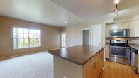Located at 11625 Community Center Drive in Northglenn, Colorado, this single-floor residence offers a well-designed open-concept layout that seamlessly connects the kitchen and living areas. The kitchen is thoughtfully equipped with stainless steel appliances, including a refrigerator, dishwasher, and range, complemented by light wood cabinetry, dark granite-style countertops, and a central island that provides additional prep and casual dining space. Track lighting overhead adds focused illumination throughout the kitchen. Hardwood-style flooring runs through the kitchen and bathroom areas, while plush carpet covers the living and bedroom spaces for added comfort. The living area is generously proportioned, bathed in natural light from large windows, and opens to a glass-panel door leading to an outdoor balcony. The bedrooms are spacious, with one featuring a vaulted ceiling, built-in shelving, and convenient closet access. A sliding glass door in another bedroom provides direct balcony access and ample natural light. The bathroom includes a full soaking tub with white tile surround accented by a gray tile border, a vanity with cabinetry, and hardwood-style flooring. Warm, neutral-toned walls throughout create a cohesive and inviting atmosphere.