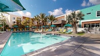 Located at 1902 Millenia Ave in Chula Vista, California, this outdoor amenity space offers a resort-style experience designed for relaxation and recreation. The centerpiece of the area is a generously sized swimming pool with crystal-clear blue water, complemented by a spa, both finished with classic blue tile detailing. Surrounding the pool is a spacious sun deck outfitted with an abundance of lounge chairs and vibrant teal umbrellas, providing ample shade and comfort for residents. Towering palm trees are strategically placed throughout the courtyard, enhancing the tropical ambiance and offering natural shade. The pool area is enclosed by a gated perimeter, ensuring privacy and security. Adjacent to the pool, a dedicated outdoor shower and restroom facility provide convenient amenities for guests. The architecture of the surrounding multi-story buildings features a contemporary design with bold color accents — including yellow, teal, and warm wood tones — along with private balconies that overlook the pool courtyard. Decorative tilework adds an artistic touch to select exterior walls. The overall atmosphere is lively yet relaxed, evoking a Southern California resort setting bathed in natural sunlight, making it an inviting outdoor retreat for the community.