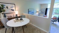 This single-floor residence offers a thoughtfully designed open-concept layout that seamlessly connects the dining and living areas, creating a cohesive and functional living environment. The dining area features a round white table accented with decorative objects, complemented by mid-century modern style chairs and warm lamp lighting that adds a welcoming ambiance. A low half-wall divider elegantly separates the dining space from the adjacent living area, maintaining an open and airy feel while providing a subtle sense of definition between zones. The living area is furnished with a comfortable sofa adorned with accent pillows, flanked by a tripod floor lamp that contributes to the room's layered lighting scheme. Throughout the space, soft neutral wall tones in muted green-gray hues provide a calm and versatile backdrop. Plush wall-to-wall carpeting adds warmth and comfort underfoot. Framed artwork is displayed thoughtfully across the walls, lending a curated, personalized character to the interior. The overall ambiance is relaxed yet refined, with a cohesive design aesthetic that balances simplicity and style, making the space feel both livable and inviting.