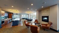 This single-floor communal space is thoughtfully designed to serve multiple functions, combining a lounge, dining area, and full kitchen into one cohesive, open-plan environment. The layout flows naturally between distinct zones, each with its own character while maintaining a unified aesthetic throughout.

The lounge area anchors one end of the space, centered around a striking floor-to-ceiling stone-clad fireplace. Cognac leather swivel chairs arranged around a glass-top coffee table create an inviting seating area ideal for casual gatherings. Adjacent to the fireplace, a separate lounge nook features a plush sectional sofa facing a large flat-screen display, with expansive floor-to-ceiling windows offering views of an outdoor terrace and surrounding greenery.

The kitchen is a functional centerpiece, featuring a bold teal island with a white quartz countertop and bar seating. Warm wood cabinetry, stainless steel appliances, a herringbone tile backsplash, and a full-size refrigerator make it well-equipped for entertaining. Overhead, a sculptural brass-and-Edison-bulb chandelier adds a contemporary industrial accent.

A dining area with long communal tables and modern chairs sits adjacent to a generous window wall, ensuring the entire space benefits from abundant natural light. Patterned carpeting throughout ties the various zones together, contributing to the space's modern yet welcoming ambiance.