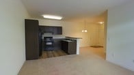 This single-floor residence offers an open-concept layout that seamlessly connects the kitchen and living areas, creating a spacious and functional environment. The kitchen is outfitted with rich, dark cabinetry that provides ample storage, complemented by matching black appliances including a gas range, refrigerator, and built-in microwave. A peninsula with a dark countertop extends from the kitchen, offering additional prep space and a natural divider between the kitchen and the adjoining living area. The sink is positioned along the peninsula, adding to the kitchen's practicality. Hardwood-style flooring anchors the kitchen space, while plush neutral carpeting flows through the living area, adding warmth and comfort underfoot. The living space is open and airy, with soft, neutral-toned walls that lend a clean and inviting ambiance. A decorative chandelier hangs near the entryway, providing an elegant touch, while recessed and flush-mount lighting ensures the space is well-lit throughout. A paneled entry door with a welcoming mat completes the foyer area. Overall, the space balances practical design with comfortable living in a cohesive, well-appointed layout.