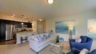 Located at 130 Ocean Aisle Circle in Salisbury, Maryland, this single-floor residence offers a thoughtfully designed open-concept layout with a warm, coastal aesthetic throughout. The living area serves as the heart of the home, featuring plush carpeting underfoot and neutral-toned walls that create a bright, inviting atmosphere. A large ocean-inspired wall mural acts as a striking focal point, reinforcing the coastal theme and adding a sense of depth to the space. The living room is comfortably appointed with ample seating and a glass-top coffee table, making it ideal for both relaxing and entertaining. The kitchen flows seamlessly from the living area, separated by a breakfast bar with seating — a practical and social feature for everyday living. Dark cabinetry contrasts elegantly with light granite countertops, while pendant lighting adds a stylish touch above the bar. Stainless steel appliances, including a range and microwave, complete the well-equipped kitchen. Recessed lighting throughout the kitchen ensures the space is both functional and visually appealing. The entry door is conveniently accessible from the main living space, contributing to the home's efficient and cohesive single-level floor plan. Overall, the space exudes a relaxed, beach-inspired charm.