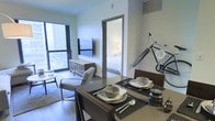 This single-floor unit is designed with an efficient, open-concept layout that seamlessly connects the dining and living areas. The dining space is positioned near the entrance and features a dark-toned table set for two, complemented by woven placemats and coordinated tableware, lending a clean and contemporary feel.

The living area flows directly from the dining space, anchored by a wall-mounted flat-screen television above a low-profile media console with a reflective finish. A pair of upholstered chairs provide seating and face toward the room's standout feature: a floor-to-ceiling window that frames expansive urban views, flooding the space with natural light and creating a strong visual connection to the city outside.

A separate bedroom is visible through an interior doorway, revealing a neatly made bed and soft lighting, suggesting a quiet and restful environment. The overall interior palette is neutral, with light-toned walls and hardwood-style flooring that enhance the sense of openness throughout. The design strikes a balance between functionality and modern aesthetics, making efficient use of the available space.