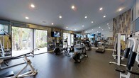 This contemporary fitness center is thoughtfully designed to support a complete workout experience. The space is outfitted with a diverse selection of cardio equipment, including treadmills, elliptical trainers, and stationary bikes, all arranged to maximize usability and flow throughout the floor. Large floor-to-ceiling windows line one side of the room, flooding the interior with natural light and offering views of lush greenery outside, creating an energizing and open atmosphere. Full-length mirrors along the walls visually expand the space and allow users to monitor their form during exercise. Recessed lighting in the smooth ceiling provides bright, even illumination throughout, complementing the natural light. Multiple flat-screen televisions are mounted at strategic points, keeping members entertained during their workouts. The dark rubberized flooring is durable and functional, suitable for both cardio and strength training activities. A dedicated free weights area is visible along the perimeter, rounding out the facility's offerings. The overall design strikes a balance between practicality and modern aesthetics, making this fitness center a well-appointed and inviting space for exercise.
