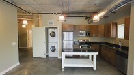 This single-floor space showcases a distinctive industrial aesthetic, characterized by exposed concrete ceilings, visible ductwork, and polished concrete floors that together create an urban loft atmosphere. The open-concept kitchen serves as a focal point, equipped with stainless steel appliances including a French door refrigerator, a gas range with microwave above, and a dishwasher. Warm-toned wood cabinetry lines the walls, providing ample storage, while pendant lights with metallic shades hang from the ceiling, casting focused illumination over the workspace. A freestanding kitchen island on casters features a dark countertop and an open lower shelf, offering additional prep space and flexibility in layout. A notable convenience is the in-unit stacked washer and dryer, neatly positioned beside a white-paneled door, maximizing utility without sacrificing floor space. The exposed HVAC ducting and raw ceiling finish reinforce the industrial character throughout, while the concrete flooring adds durability and visual continuity. The overall ambiance is functional yet stylish, blending practical amenities with a bold, urban design sensibility.