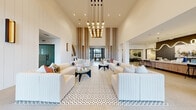 Located along Tamiami Trail East in Naples, Florida, this single-story clubhouse offers a refined and thoughtfully designed gathering space suited for community socializing and recreation. The grand central lounge immediately commands attention with its dramatically soaring ceilings adorned by a cascading cluster chandelier with exposed globe bulbs. Warm wood vertical slat panels frame the corridor leading to expansive floor-to-ceiling windows at the far end, drawing natural light deep into the interior. Geometric patterned tile flooring anchors the seating area, which is furnished with plush cream sofas and sculptural dark-toned accent tables.

An adjacent game and entertainment room features large-format neutral tile flooring, a pool table, mounted flat-screen television, and bold circular wall art against textured paneling. A communal kitchen area offers a generous white-surface island with bar seating, dark tile backsplash, wood cabinetry, and dual wall-mounted screens. Delicate wood slat screens elegantly partition one section of the space, adding visual texture and depth.

A cozy café nook, complete with a coffee station and light wood cabinetry, opens to floor-to-ceiling black-framed windows overlooking lush Florida landscaping, creating an inviting, sun-drenched retreat within the clubhouse.
