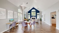 This single-floor leasing office is defined by its striking architectural design and welcoming atmosphere. The centerpiece of the space is a dramatic vaulted ceiling that rises to impressive heights, drawing the eye upward toward a bold navy blue accent wall adorned with an arched transom window and flanked by large pane windows that flood the interior with natural light. A classic white fireplace anchors the lounge area, which features tufted armchairs arranged around a round accent table atop a patterned area rug — ideal for relaxed client consultations. Rich wood-look plank flooring runs throughout, providing a warm, cohesive foundation across all areas. A casual meeting zone near the entrance offers a marble-top round table surrounded by modern upholstered chairs in muted lavender, positioned beneath the soaring ceiling peak. French doors along the front façade open to views of lush greenery outside, reinforcing the bright and airy feel. A dedicated workstation area provides a professional, functional workspace with built-in cabinetry and multiple computer stations. Soft gray walls complement the navy accents, creating a polished yet approachable environment. The overall ambiance is modern and sophisticated, thoughtfully designed to welcome and inform prospective residents.