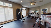 This single-level fitness space is thoughtfully designed to support a wide range of workout routines. The room features neutral taupe walls, recessed lighting, and ceiling fans that contribute to a comfortable and functional exercise environment. Rubber flooring covers the entire area, providing both durability and safety underfoot.

Natural light enters generously through multiple large windows fitted with white horizontal blinds, including transom windows positioned above the main panes. These windows are distributed across several walls, ensuring the space feels bright and open. Strategic placement of large wall-mounted mirrors further amplifies the sense of space and allows users to monitor their form during exercise.

The cardio zone is well-appointed with multiple treadmills and an elliptical trainer, each equipped with individual monitors. Strength training is supported by a multi-function cable machine, a dedicated free-weight rack stocked with a full set of dumbbells, a weight bench, and additional resistance equipment including a rowing-style machine and stationary bike.

The overall layout is organized to allow clear movement between equipment zones. Ventilation is supported by ceiling fans positioned throughout the room, and informational signage is posted on the walls, reinforcing the space's professional fitness-center character.