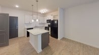 This single-floor residence is thoughtfully designed with a cohesive, contemporary aesthetic carried throughout its living spaces. Light-toned hardwood-style flooring flows consistently across the main areas, creating a unified and airy feel.

The kitchen serves as a functional centerpiece, outfitted with white shaker-style cabinetry accented by brushed nickel hardware and granite-look countertops. A classic white subway tile backsplash adds visual texture, while black appliances — including a range, over-the-range microwave, dishwasher, and refrigerator — provide a bold contrast. A dark-based kitchen island with a granite surface offers additional prep space and is illuminated by pendant lighting above, complemented by a dedicated wine rack built into the cabinetry nearby.

The living area is generously proportioned, featuring a ceiling fan with integrated lighting and a set of French doors that invite natural light and provide access to the outdoors. The open layout connects seamlessly to the kitchen and adjacent carpeted space.

The bathroom is finished with a white vanity, granite countertop, large mirror with overhead lighting, and a tub-shower combination with tile surround. Chrome fixtures and a towel bar complete the clean, practical design.