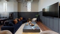 Located at 538-546 Broad Street in Newark, NJ, this single-floor lounge is a thoughtfully designed communal space that blends refined aesthetics with practical functionality. Rich hardwood flooring flows throughout, unifying the various zones within the space.

The main lounge area makes a striking impression with soaring ceilings, bold charcoal board-and-batten wainscoting, and dramatic tall windows featuring chevron-patterned glazing that flood the room with natural light. Deep navy sofas, leather-accented pillows, and a substantial stone coffee table anchor the seating area beneath a geometric chandelier, while a large wall-mounted display screen adds a modern touch.

The fully equipped kitchen features an expansive dark stone-topped island with bar seating, stainless steel appliances, and open shelving accented by white subway tile. Globe pendant lights overhead complete the polished yet welcoming atmosphere.

A dedicated private conference room offers a long dark wood table surrounded by upholstered chairs, a warm walnut feature wall, and an artistic multi-arm chandelier — ideal for focused meetings or private gatherings.

Near a wide staircase, a casual seating nook with bar stools and a classic billiards cue cabinet adds a relaxed, social dimension. Throughout, the design balances contemporary sophistication with inviting warmth, making this lounge a versatile and well-appointed destination.