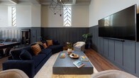 Located at 538-546 Broad Street in Newark, NJ, this single-floor lounge is a thoughtfully designed communal space that blends refined aesthetics with practical functionality. Rich hardwood flooring flows throughout, unifying the various zones within the space.

The main lounge area makes a striking impression with soaring ceilings, bold charcoal board-and-batten wainscoting, and dramatic tall windows featuring chevron-patterned glazing that flood the room with natural light. Deep navy sofas, leather-accented pillows, and a substantial stone coffee table anchor the seating area beneath a geometric chandelier, while a large wall-mounted display screen adds a modern touch.

The fully equipped kitchen features an expansive dark stone-topped island with bar seating, stainless steel appliances, and open shelving accented by white subway tile. Globe pendant lights overhead complete the polished yet welcoming atmosphere.

A dedicated private conference room offers a long dark wood table surrounded by upholstered chairs, a warm walnut feature wall, and an artistic multi-arm chandelier — ideal for focused meetings or private gatherings.

Near a wide staircase, a casual seating nook with bar stools and a classic billiards cue cabinet adds a relaxed, social dimension. Throughout, the design balances contemporary sophistication with inviting warmth, making this lounge a versatile and well-appointed destination.