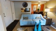 This single-story space showcases an open-concept design that seamlessly connects the living, dining, and kitchen areas into one cohesive environment. The living area is anchored by a light blue sofa adorned with patterned accent pillows, complemented by a decorative sunburst wall piece and a pass-through window that provides a direct sightline into the kitchen. The kitchen features a countertop bar area visible through the pass-through, offering both functionality and visual connectivity to the main living space. A striking accent wall in deep orange defines the dining area, which is equipped with a round table and dark dining chairs beneath a ceiling fan with integrated lighting. Framed floral artwork decorates the dining walls, adding a curated aesthetic to the space. The flooring transitions between carpet in the living zone and harder surfaces near the dining area. Teal decorative vases on the coffee table introduce a vibrant pop of color that ties together the blue and orange palette throughout. The overall ambiance is warm, bold, and inviting, with a well-considered use of color and decor to define each functional zone within the open layout.
