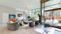 The lobby at Park Fifth is a striking, contemporary space designed to make a lasting first impression. Soaring ceilings amplify the sense of grandeur, while expansive floor-to-ceiling glass walls flood the interior with abundant natural light and offer unobstructed views of the surrounding streetscape and greenery. Large-format porcelain tile flooring in a soft neutral tone extends throughout, lending a clean and polished foundation to the space.

At the heart of the lobby, a generous seating area features an eclectic mix of curved and tufted furniture in muted blues, grays, and purples, all anchored by a round area rug. Cascading tiered chandeliers with delicate fringe detailing hang overhead, adding an elegant focal point. Curated artwork adorns the textured accent walls, contributing warmth and visual interest.

Adjacent to the main seating area, a dedicated concierge desk with a distinctive marble-fronted panel and warm gold accents serves as the welcome point for residents and visitors. A nearby corridor reveals a set of floor-to-ceiling operable partition panels, providing flexible use of space for events or gatherings. Lush indoor plants are thoughtfully integrated throughout, enhancing the connection between the interior and the vibrant outdoor environment visible through the generous glazing.