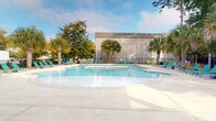 Located at 206 Cedar Branch in Wilmington, North Carolina, this single-level outdoor amenity space offers a well-appointed, resort-inspired environment designed for relaxation and recreation. At its centerpiece is a generously sized, freeform swimming pool with crystal-clear blue water, surrounded by an expansive concrete sundeck lined with teal lounge chairs — ideal for soaking up the Carolina sunshine. The pool area is framed by a wrought iron perimeter fence and accented by mature palm trees and established shade trees, giving the space a distinctly coastal, tropical atmosphere. A dedicated lounge zone features wicker seating with colorful striped cushions, shaded by a large market umbrella and anchored by a prominent palm tree, creating an inviting retreat within the deck. A covered pavilion with a ceiling fan provides a shaded gathering area and houses a ping-pong table, offering a fun recreational option just steps from the pool. Stone-clad columns lend architectural character to the pavilion structure, which also features white-trimmed double-entry doors with large windows, connecting seamlessly to an interior space beyond. The overall ambiance is bright, open, and relaxed, capturing the laid-back lifestyle of coastal Wilmington.