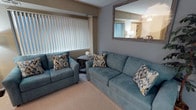 This single-floor unit presents a thoughtfully designed and cohesively decorated interior. The living area serves as the central gathering space, furnished with a matching teal sofa and loveseat accented by ikat-patterned throw pillows in blue, gold, and grey tones. Vertical blinds along the windows allow for adjustable natural light throughout the space. A wall-mounted flat-screen television is paired with a rustic wood media console, while a contemporary floor lamp with an illuminated column base provides warm ambient lighting.

A large framed mirror mounted on the accent wall in a blue-grey hue adds depth to the living area, visually expanding the space. Artwork featuring a serene woodland scene in soft blues and greens decorates an adjacent wall, complementing the overall color palette. The entryway transitions seamlessly from wood-look flooring into the carpeted living area, and a decorative indoor tree near the front door adds a natural touch.

The dining area is visible from the living room, featuring a dark wood table and chairs, and a glimpse into the kitchen reveals light cabinetry. Neutral warm-toned walls throughout create a calm, inviting ambiance.