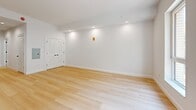 This thoughtfully designed studio unit at 229 Willett Ave in Port Chester, NY offers a clean, modern aesthetic across a single floor. The open living area is bathed in natural light from dual windows that frame views of surrounding greenery, while light-toned hardwood flooring and crisp white walls create a bright, airy atmosphere. Recessed lighting throughout enhances the sense of openness. A louvered double-door closet provides practical storage within the living space. The kitchen is well-appointed with white shaker-style cabinetry, black hardware accents, a white quartz countertop, a deep single-basin sink with a matte black faucet, a gray tile backsplash, and stainless steel appliances including a full-size refrigerator. A warm wood-toned entry door anchors the kitchen area. The bathroom showcases a refined, contemporary design with floor-to-ceiling stone-look wall panels in a neutral beige tone, a walk-in shower with a matte black showerhead, a sleek white floating vanity with an undermount sink, a matte black faucet, and a large backlit mirror. Together, these elements deliver a cohesive, move-in-ready space with modern finishes at every turn.