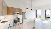 Located on the fifth floor at 282 Bremen Street in Boston's East Boston neighborhood, Unit 507 is a thoughtfully designed one-bedroom, one-bathroom residence that blends contemporary aesthetics with functional living. The open-concept layout seamlessly connects the kitchen and living area, creating an airy, cohesive space enhanced by soaring ceilings that amplify the sense of openness throughout.

The kitchen is a standout feature, showcasing a clean design with white lower cabinetry, warm wood-toned upper cabinets, stainless steel appliances including a full-size refrigerator, range, microwave, and dishwasher, and a white quartz-topped island ideal for dining or meal prep. Globe pendant lights add a modern touch overhead. Light hardwood flooring flows continuously through the main living areas, complementing the bright white walls.

The bedroom is generously proportioned and bathed in natural light through floor-to-ceiling black-framed windows that frame verdant outdoor views. The bathroom features a soaking tub with crisp white subway-style tile surround, maintaining the unit's clean, minimalist aesthetic. A dedicated closet provides practical storage. Throughout, the unit's elevated position offers scenic treetop and skyline views, making it a bright and inviting urban retreat.