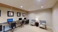 This single-floor business center is thoughtfully designed to provide a functional yet comfortable workspace. The room features a sleek granite-topped workstation along one wall, equipped with multiple desktop computers and ergonomic adjustable office chairs, making it well-suited for focused work or research tasks. Recessed lighting is installed throughout the ceiling, ensuring consistent, even illumination across the entire space. The neutral beige walls and light-toned flooring contribute to a clean, professional ambiance. A dedicated lounge area complements the workstation zone, furnished with cream-colored accent chairs adorned with bold black-and-white checkered cushions, paired with a round dark wood side table and a classic floor lamp that adds a warm glow. Framed artwork in dark borders decorates the walls, adding visual interest and a refined touch to the overall aesthetic. The layout strikes a balance between practicality and comfort, offering both a productive desk environment and a relaxed seating area within the same space. The design is cohesive and modern, making this business center an efficient and inviting resource for those seeking a dedicated place to work.