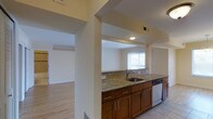 This single-story, one-bedroom, one-bathroom residence offers a thoughtfully designed layout that balances functionality and comfort. The kitchen serves as a central feature of the home, showcasing warm medium-toned wood cabinetry paired with granite countertops that provide generous prep space. A stainless steel sink and dishwasher add a modern, practical touch, while a classic tile backsplash introduces subtle texture to the space. The kitchen opens toward the adjacent living area through a structural column framing, creating a sense of visual connectivity between the two spaces. Light tile flooring runs through the kitchen, transitioning to warm hardwood-style flooring in the living areas, adding a cohesive flow throughout the home. Recessed lighting provides even illumination in the kitchen, while large windows dressed with vertical blinds allow natural light to filter into the living space. The neutral, light-toned walls throughout contribute to a bright and airy ambiance. Overall, the residence presents a clean, move-in-ready aesthetic with durable finishes well-suited for everyday living.